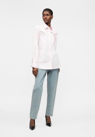 Femme debout portant une chemise rose pâle à manches longues boutonnée, un jean droit bleu clair et des talons pointus noirs sur fond blanc.