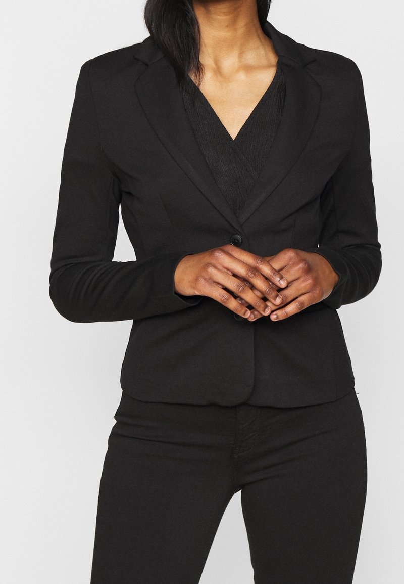 Blazer noir avec une fermeture à un seul bouton, coupe cintrée, revers crantés et manches longues ; porté sur un haut texturé noir.