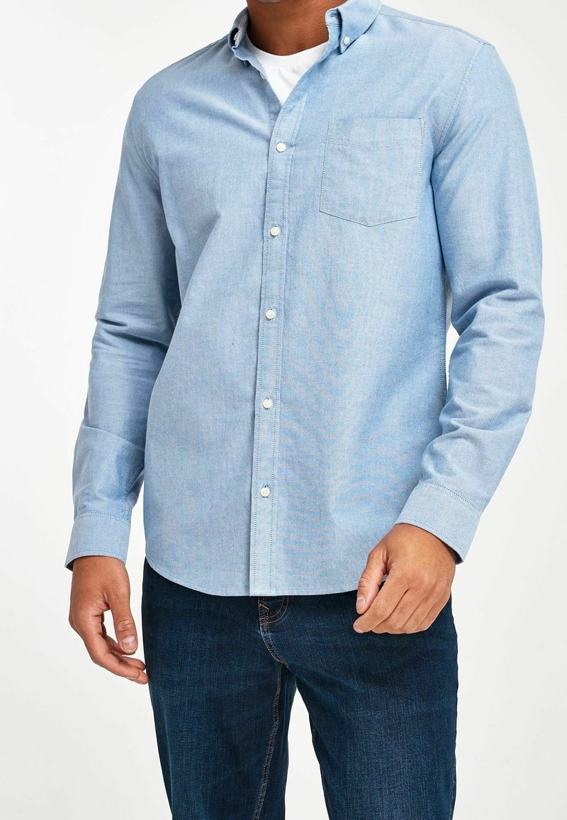 Homme portant une chemise bleu clair à boutons sur un sous-vêtement blanc et un jean bleu foncé, debout, les mains près des hanches sur fond blanc.