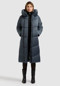 Prešívaná bunda tmavomodrej farby s vysokým golierom, dlhými rukávmi a predným zipsom. Má hladký, lesklý povrch a siaha až po kolená.