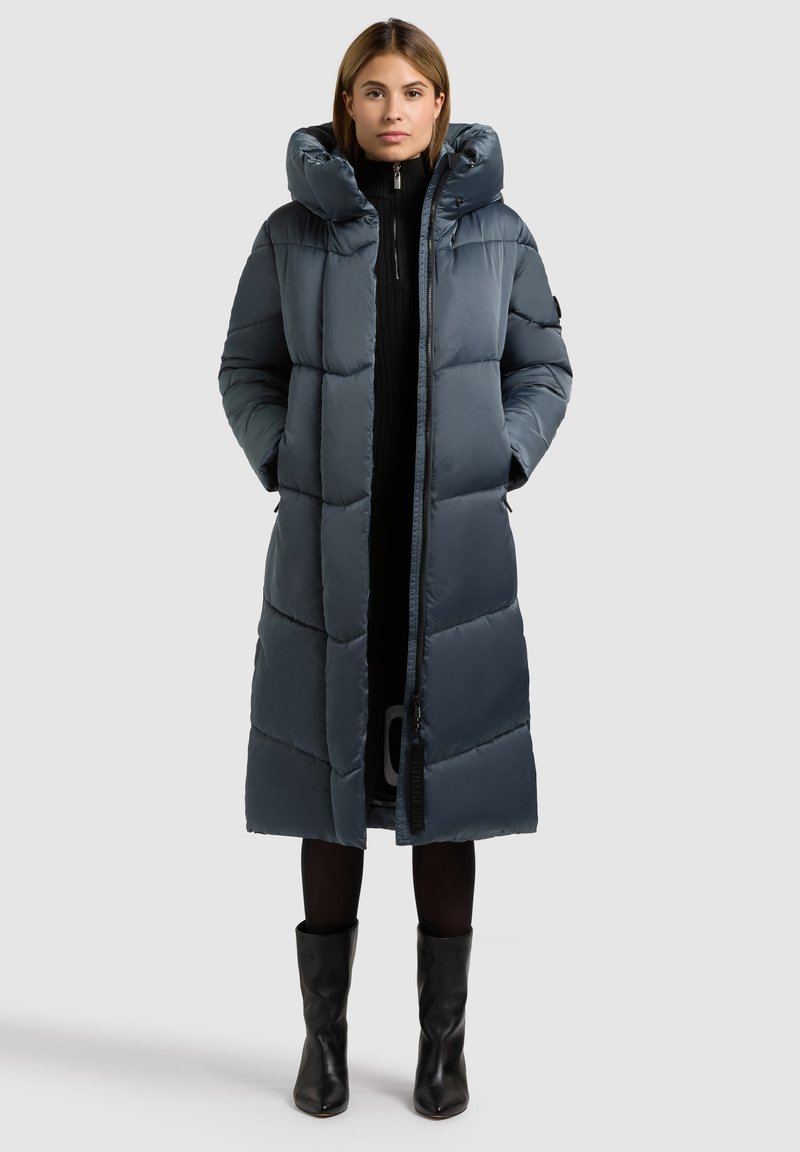 Sötétkék steppelt kabát, magas gallérral, hosszú ujjakkal és elülső cipzárral. Simább, fényes textúrájú, és térdig érő hosszúságú.