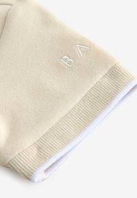 Manica in tessuto beige lavorato a maglia con polsino a coste e lettere ricamate bianche "BA" vicino al bordo su sfondo bianco.