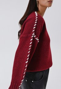 Donna che indossa un maglione a maglia bordeaux con dettagli in filato bianco attorcigliato lungo le maniche, abbinato a jeans di denim neri.
