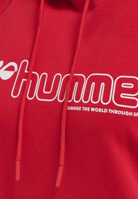 Czerwona bluza z kapturem z białym tekstem graficznym. Posiada sznurki z czerwonymi końcówkami. Materiał wydaje się miękki i teksturowany, odpowiedni do noszenia na co dzień.