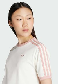 Hvit kortermet t-skjorte med rosa detaljer, har rund hals, teksturerte striper på skuldrene, og et brodert logo på brystet.