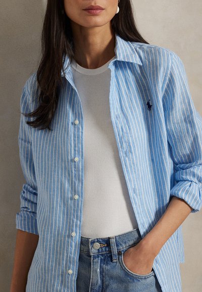 Femeie purtând o cămașă cu dungi albastre deschise și albe, nasturată, peste un top alb cu textură reiată, cu mâna în buzunarul blugilor albaștri.