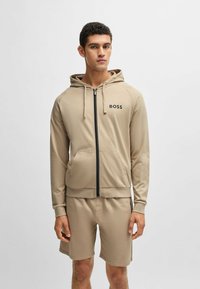 Sudadera beige con cremallera y capucha, bolsillos frontales y logo negro de "BOSS". Combinada con pantalones cortos beige a juego, hechos de una tela suave.
