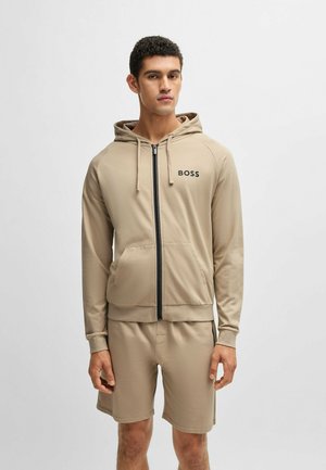 Haut à zip beige avec une capuche, poches avant et logo noir "BOSS". Porté avec un short beige assorti, fabriqué en tissu lisse.