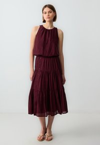 Bordeauxrotes, ärmelloses Kleid mit gerafftem Ausschnitt, gestuftem Design und Knopfdetails an der Seite, aus leichtem Stoff gefertigt.