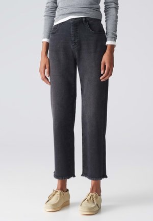 Person trägt hoch taillierte, gerade geschnittene schwarze Jeans mit ausgefransten Säumen, graues Langarmshirt und beige Schnürschuhe, steht vor schlichter Hintergrund.