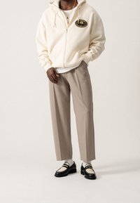 Crèmekleurige hoodie met ritssluiting en logo, beige broek met brede pijpen en zwarte schoenen met crèmekleurige accenten. Gestyled met witte sokken eronder.