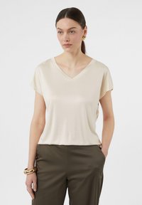 Maglietta beige a V con maniche corte, texture morbida e vestibilità rilassata, abbinata a pantaloni verde oliva. Design semplice e versatile.
