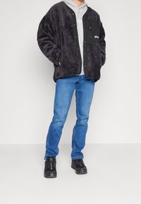 Persona che indossa una giacca in pile nera sopra un felpa grigia chiara, jeans blu e sneakers nere, in piedi con le mani nelle tasche della giacca.
