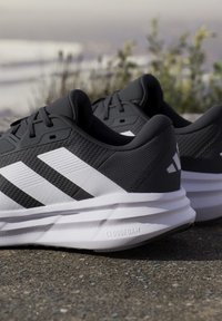 Paire de chaussures de course Adidas noires et blanches avec semelles Cloudfoam posées sur l'asphalte près de verdure et d’un arrière-plan d’eau flou.