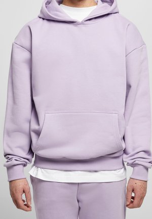 Person trägt einen hellvioletten Hoodie mit Kängurutasche und passende Jogginghose, darüber ein weißes T-Shirt.