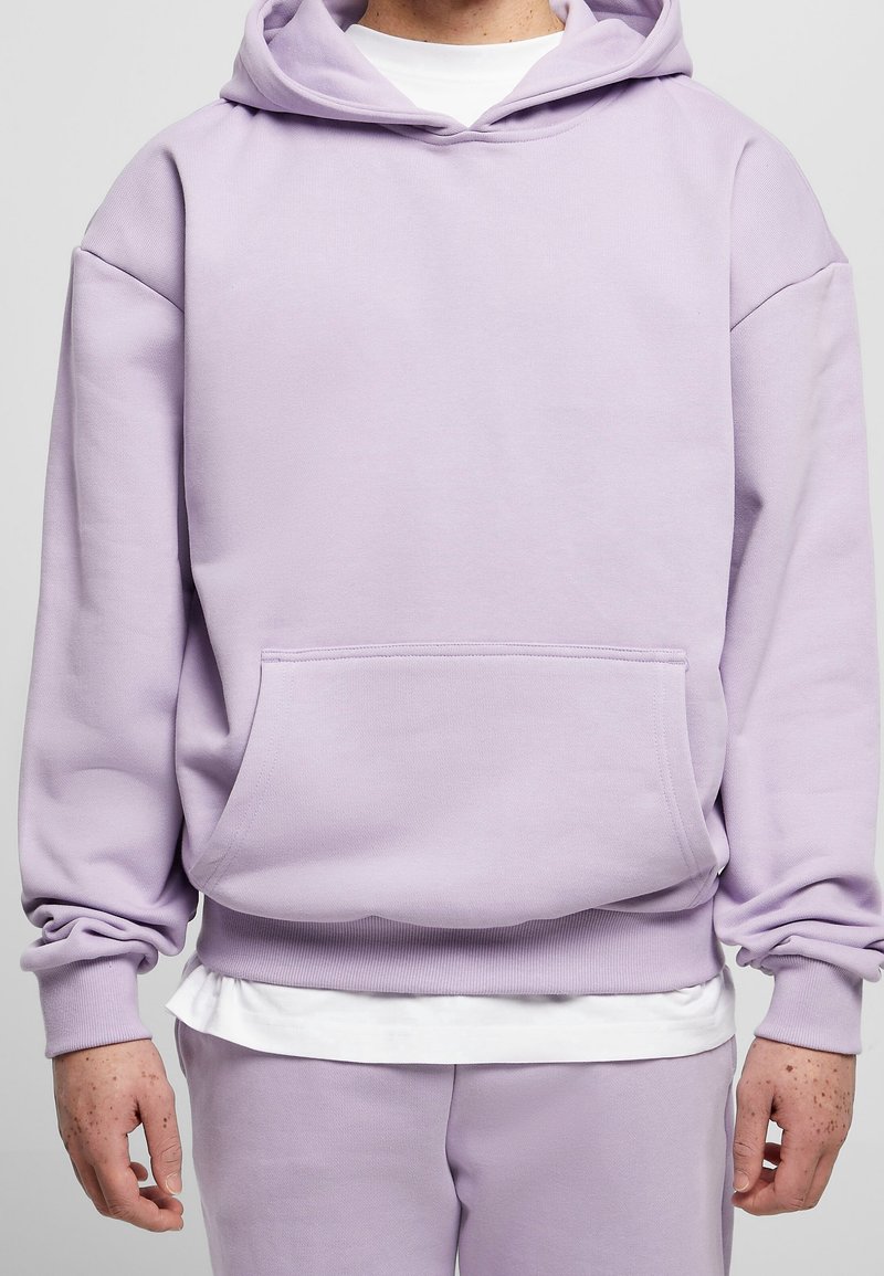 Personne portant un sweat à capuche violet clair avec une poche kangourou et un pantalon de jogging assorti, superposé à un t-shirt blanc.