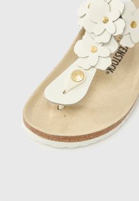 Λευκό σανδάλι με ανθισμένες διακοσμήσεις, χρυσά μεταλλικά στοιχεία, μαλακό πάτο και σόλα φελλού. Ορατή υφή στην εσωτερική επιφάνεια και απαλές υφές.