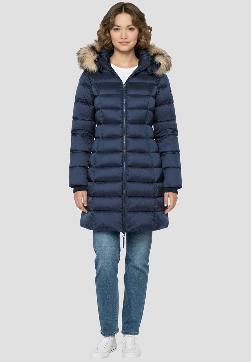 Manteau matelassé bleu marine avec une capuche doublée de fausse fourrure, fermeture éclair et motif matelassé horizontal. Porté avec un jean bleu clair et des baskets blanches.
