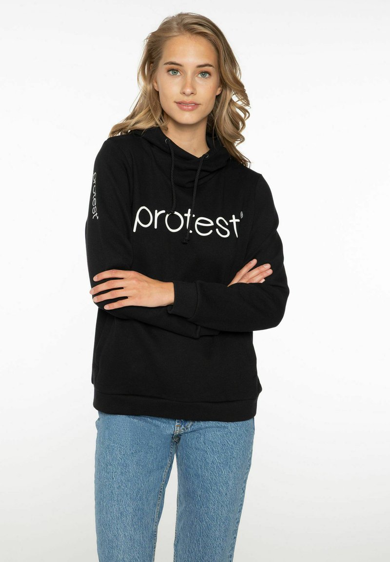 Sudadera negra con bolsillo canguro, capucha con cordón y logotipo blanco de "protesta". Combinada con jeans de color azul claro. Diseño simple y casual.
