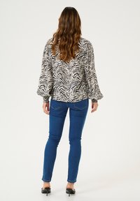 Langärmlige Bluse im Zebra-Print, beige und schwarz. Verfügt über voluminöse Ärmel mit elastischen Bündchen. Getragen mit eng anliegenden blauen Jeans und schwarzen High Heels.