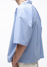 Koszula z krótkim rękawem w niebieskie paski z kołnierzykiem na guziki. Charakteryzuje się pionowymi paskami i luźnym krojem z bocznymi zaszewkami.