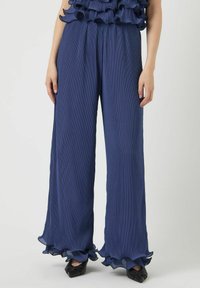 Pantalons large couleur bleu marine avec une texture plissée et un ourlet froufrou. Taille fuselée et tissu doux. Associés à des escarpins noirs à bout pointu.