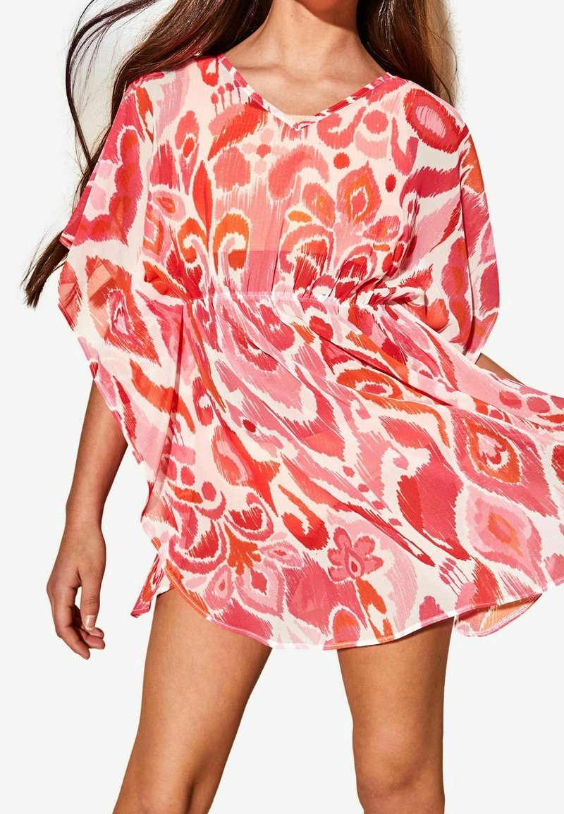 Femme portant une robe ample à motifs roses et rouges avec de larges manches, tenant la jupe légèrement vers l'extérieur.