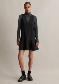 Μαύρο φόρεμα χωρίς μανίκια με V-λαιμόκοψη και φουσκωτό φούστα, συνδυασμένο με σκούρο μακρυμάνικο τοπ και παχιές μαύρες loafers. Λείο υφή.