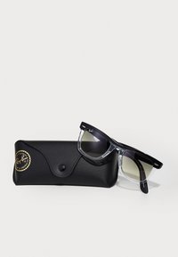 Musta ja kirkas Ray-Ban Wayfarer -aurinkolasit, joissa on ylimääräiset sävykkäät linssit, nojaamassa mustaan, teksturoituun koteloon, joka on koristeltu kultaisella Ray-Ban-logotarralla.