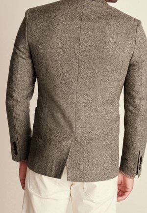 Man draagt een bruine blazer met visgraatmotief en zwarte knopen, gecombineerd met off-whitige broek, gezien van achteren.