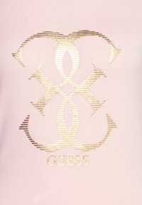 Ljusrosa t-shirt med en metallisk guldig logotypdesign med horisontella ränder och texten "GUESS" centralt placerad under logotypen.