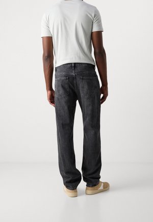 Homme portant un jean droit noir, un t-shirt blanc et des baskets beiges, debout dos à la caméra sur un fond uni.