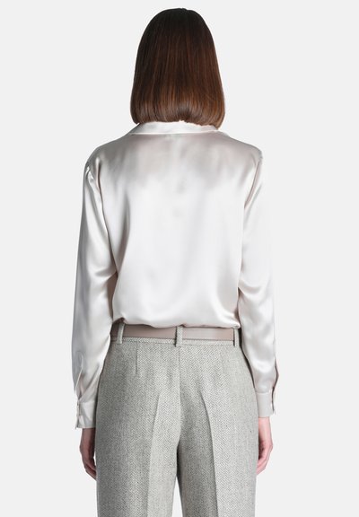 Personne aux cheveux bruns mi-longs porte un chemisier satiné crème brillant rentré dans un pantalon gris clair texturé, debout, dos face à l’avant.