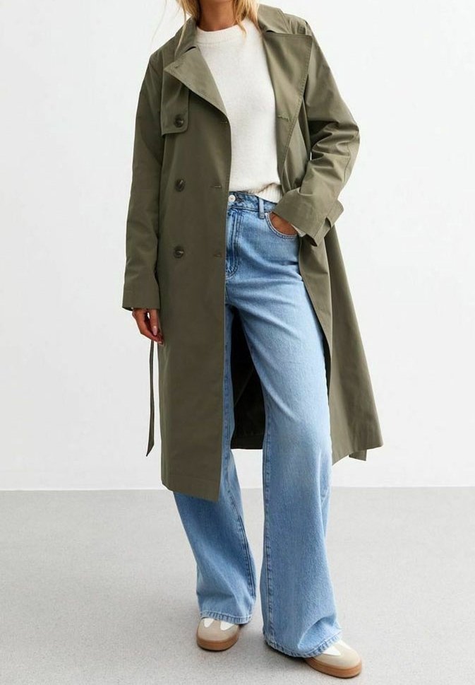 Personne portant un long trench-coat vert olive, un pull blanc, un jean large bleu clair et des chaussures couleur crème, debout avec une main dans la poche.