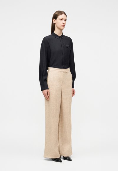 Γυναίκα όρθια φοράει μακρυμάνικο μαύρο πουκάμισο που έχει μπει μέσα σε μπεζ παντελόνι με φαρδιά μπατζάκια και ψηλή μέση, με μαύρα μυτερά παπούτσια.