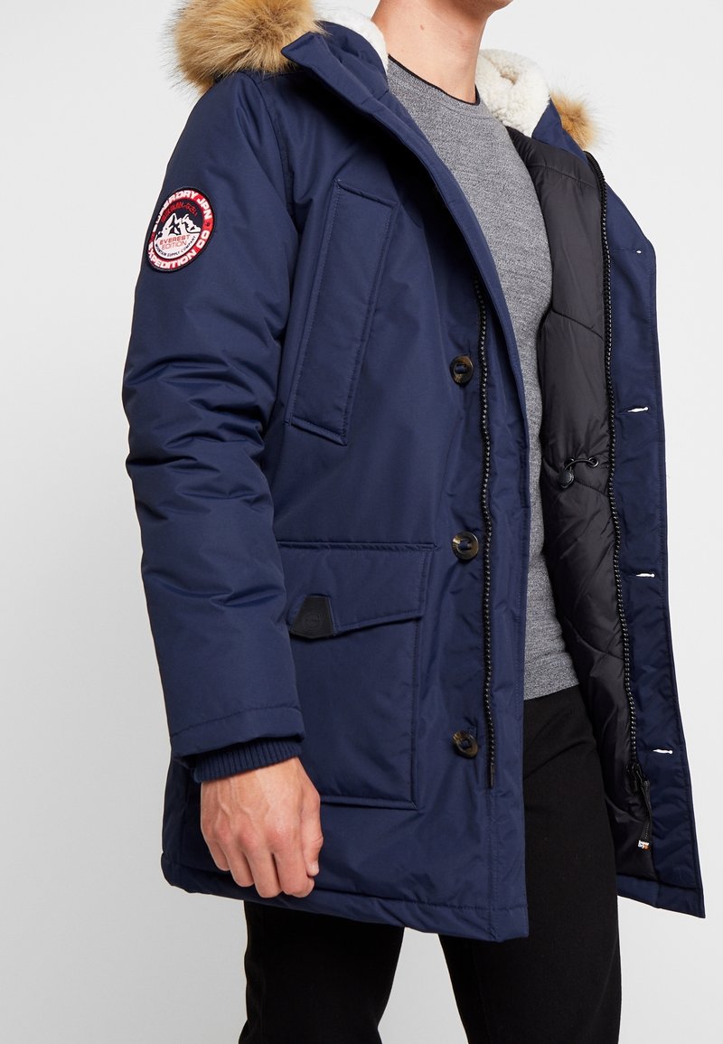 Parka bleu marine avec capuche bordée de fourrure, fermetures à boutons, multiples poches, et un écusson sur la manche. Intérieur doux et matelassé.