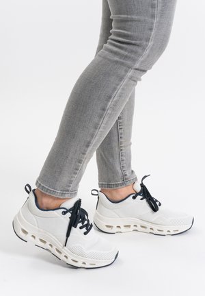 Weiße Mesh-Sneaker mit navyfarbenen Akzenten, strukturiertem Obermaterial und charakteristischem Ausschnittsohlen-Design; kombiniert mit grauen eng anliegenden Jeans.