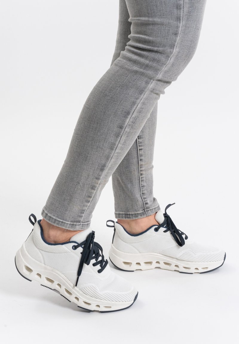 Weiße Mesh-Sneaker mit navyfarbenen Akzenten, strukturiertem Obermaterial und charakteristischem Ausschnittsohlen-Design; kombiniert mit grauen eng anliegenden Jeans.