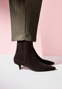 Tumši brūni zamšādas potīšu zābaki ar pīlpata purngaliem un mazu papēdi, apvienoti ar svītrainiem biksēm uz rozā fona.