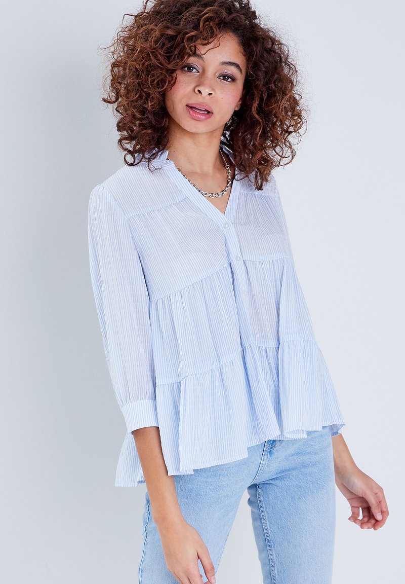 Blouse rayée bleu clair avec un design évasé en couches, col et boutonnage à l'avant. Manches longues avec poignets ; tissu texturé respirant.
