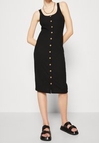 Czarna, prążkowana sukienka midi z okrągłym dekoltem i guzikami z przodu, z dwoma bocznymi kieszeniami. W zestawie z czarnymi płaskimi sandałami.