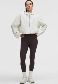 Young woman standing with hands on hips wearing white zip-up hoodie, black leggings, white socks, and white sneakers on plain background.