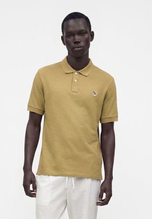 Homme portant un polo jaune moutarde à manches courtes avec un petit patch zèbre sur la poitrine et un pantalon blanc, debout devant un fond uni.