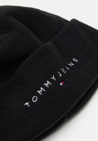 Mustat fleece-pipo, jossa on brodeerattu "TOMMY JEANS" -logo ja pieni punainen, valkoinen ja sininen yksityiskohta. Pehmeä rakenne, yksinkertainen muotoilu.