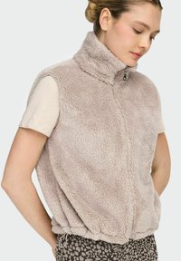 Light beige fleece vest with high collar, front zipper, and sleeveless design. Textured surface, relaxed fit, and gathered hem detail.
