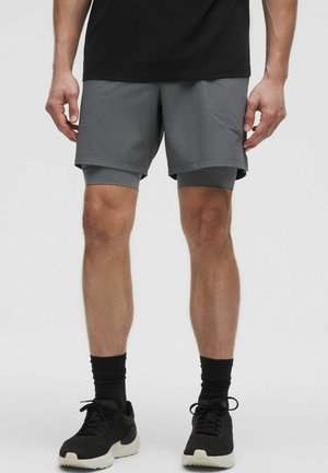 Une personne se tient debout, portant un t-shirt noir, un short de sport gris et des baskets noires. L'arrière-plan est uni et clair.