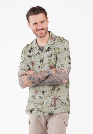 Hombre con los brazos cruzados, vistiendo una camisa verde con estampado tropical y pantalones beiges, sonriendo y mirando ligeramente de lado.
