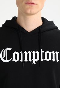 Felpa nera con cappuccio, caratterizzata da un testo bianco in stile gotico "Compton" ben in vista sul davanti. Sono visibili i lacci e una texture morbida.