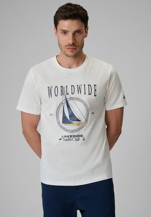 Mężczyzna z krótkimi, ciemnymi włosami ubrany w biały t-shirt z żaglówką i napisem "Worldwide Lakeside Yacht Club" oraz granatowe spodnie.