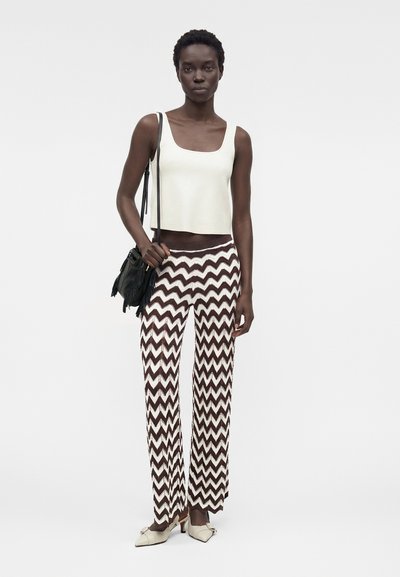 Γυναίκα με λευκό αμάνικο τοπ, καφέ και λευκό παντελόνι με ζιγκ-ζαγκ μοτίβο, λευκά παπούτσια με χαμηλό τακούνι και κρατώντας μαύρη τσάντα ώμου.
